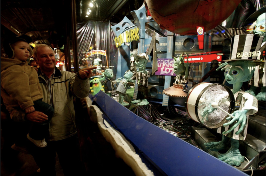 An older man holds a small boy in his arms. The man stands beside the Fenwick Christmas window and points at an alien playing a drum. This was part of the 2002 Christmas window display at Fenwick Newcastle.