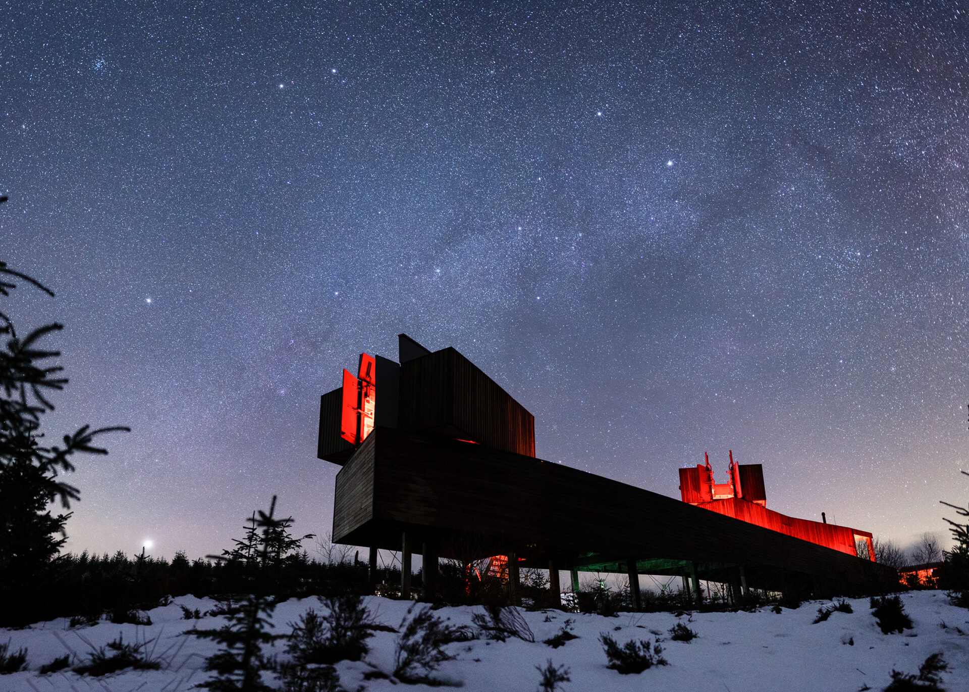 A long observatory stands in a snow covered field. Red lights illuminate it. The sky above is full of stars.