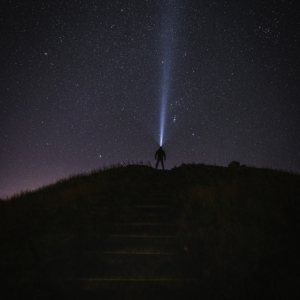 The small silhouette of a man can be seen standing at the top of a hill. He wears a head torch and looks up at the constellations in the night sky, the beam of light shining upwards. Photo by Jim Scott.