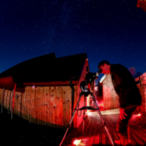 A man peers into a telescope on a wooden decking area at Battlesteads Dark Sky Observatory. The night sky above is deep blue.