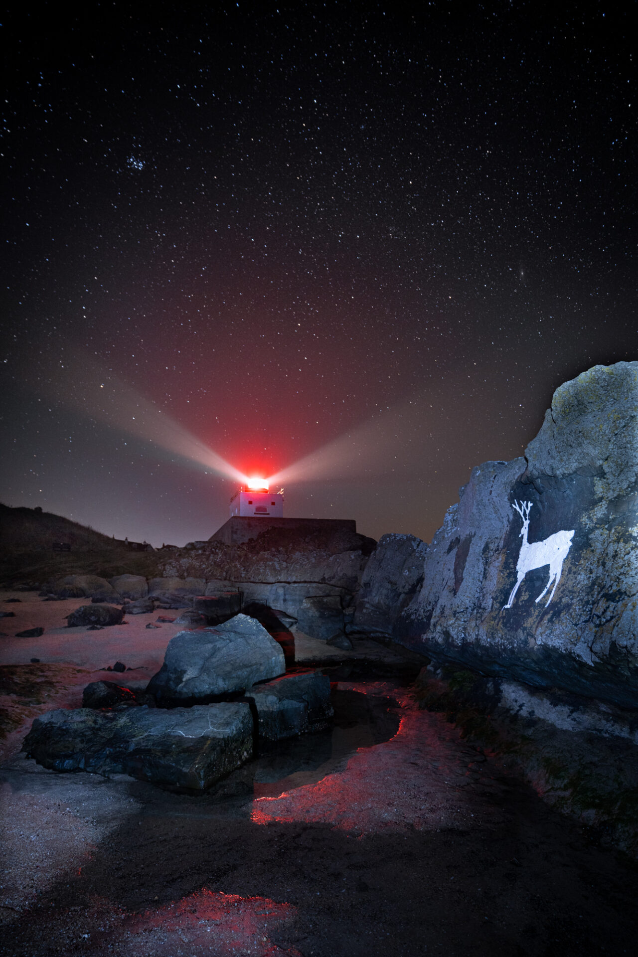A white stag is drawn onto a rock. The night sky above is illuminated by a red beacon shining from a lighthouse style building.