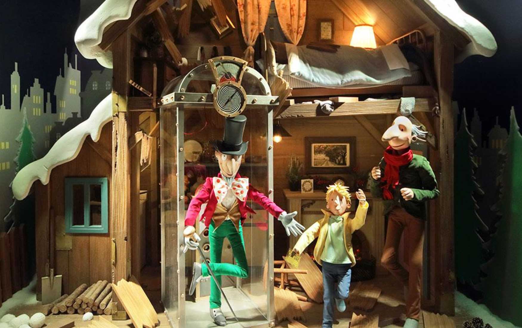 Quentin Blake style figures of Willy Wonka, Charlie and Grandpa Joe. They stand in a ramshackle house with the roof caved in, beneath which is a glass elevator. This was the Christmas window display at Fenwick Newcastle in 2019.