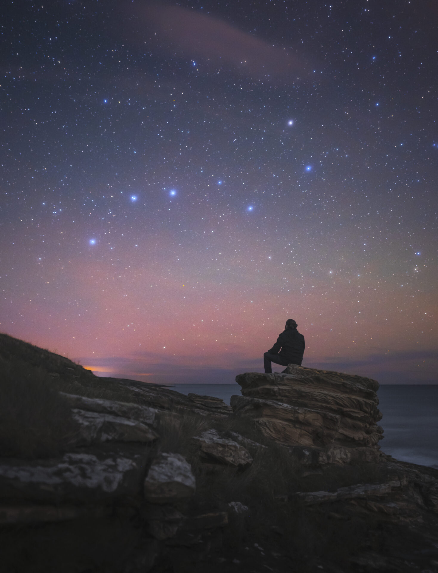 A silhouetted person sits on top of a rock in Northumberland. They gaze up at the starry sky, which is pink, purple and blue. Photo by Dan Monk.