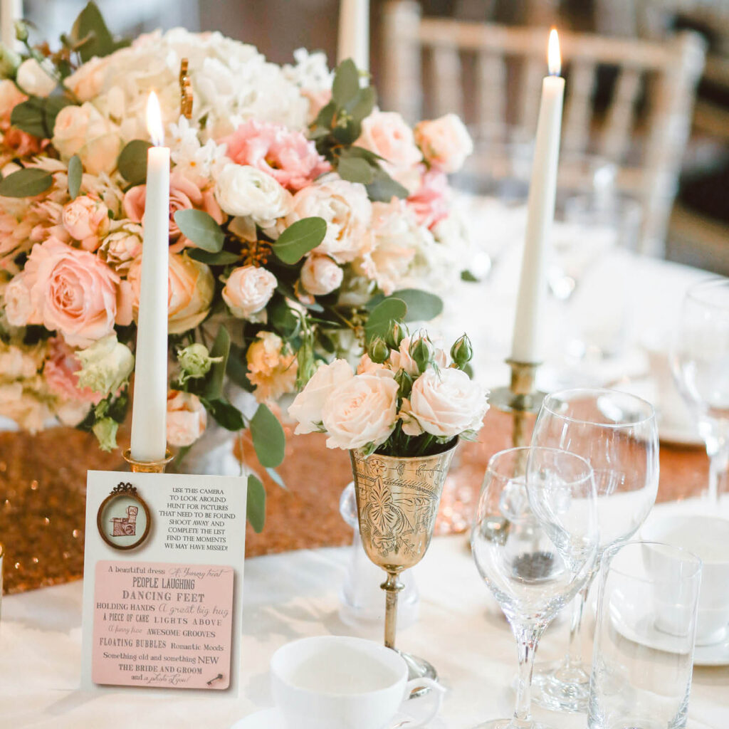Pink and white roses, tall candles and wine glasses cover a table ready for a wedding dinner.