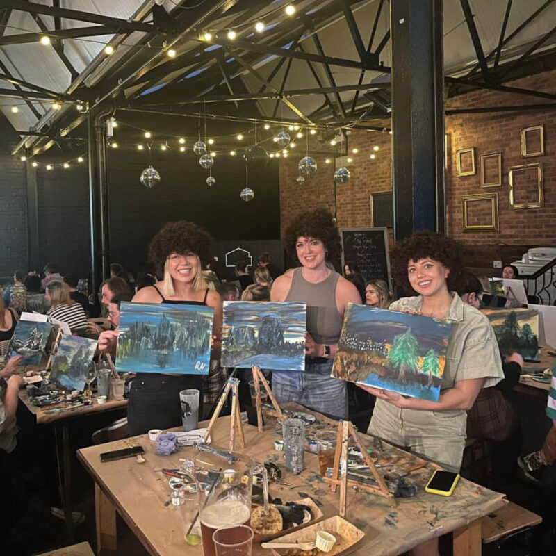 Three women stand at a table in Tyne Bank Brewery. The wear curly wigs like Bob Ross's iconic hair. They each hold up a landscape painting that they have done at the Joy of Painting evening.