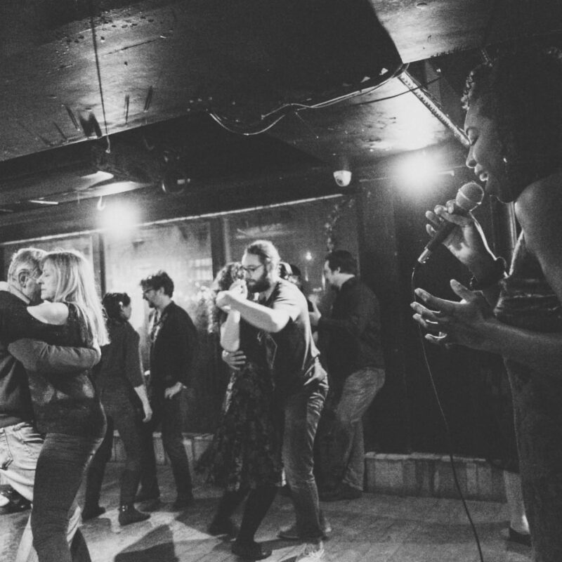 Black and white photo of a Blues dance class and social at Prohibition Cabaret Bar. A woman sings into a microphone in one corner, whilst couples hold each others hands and dance.