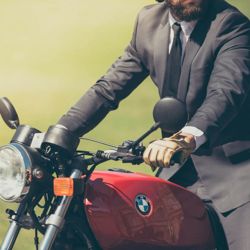 A gentleman dresses in a grey suit and tie sits astride a red BMW motorbike. His hand is poised over one handle.