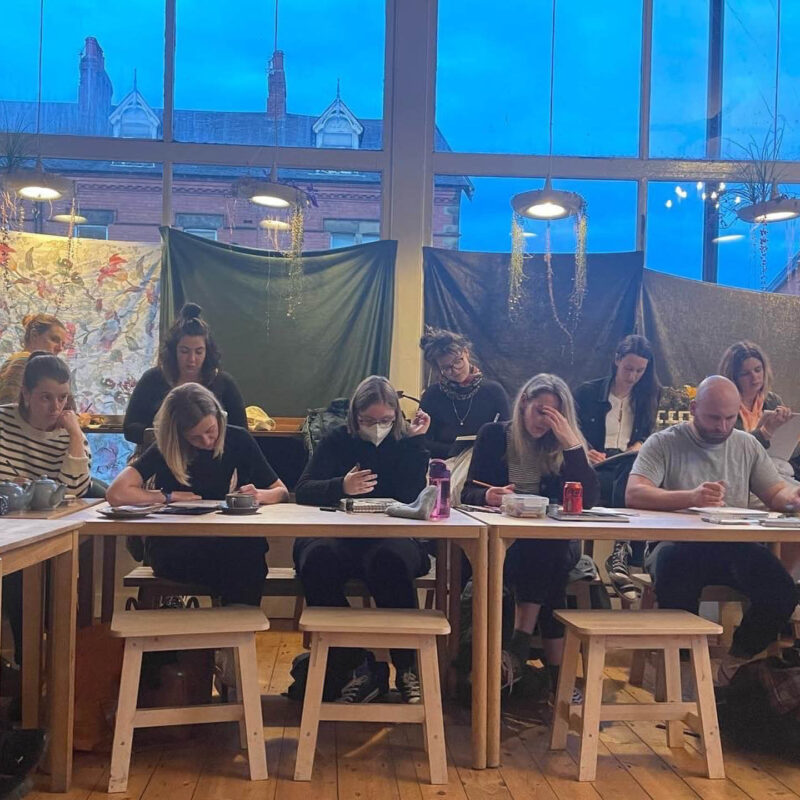 Several people sit at a table in Shanti Bee studios. They all look down at their paper in concentration as they try out life drawing.