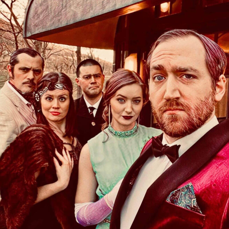 The cast behind Murder on the Disorient Express stand outside an old train carriage. They wear old fashioned suits and evening dresses. They all look look at the camera with different expressions of suspicion and disdain.