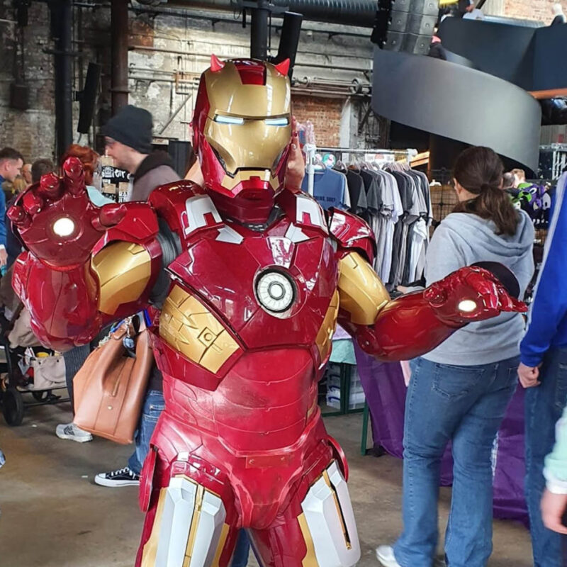 A person dressed in an Iron Man costume raises their hands towards the camera. Behind them, people browse the stalls of Nerd Fest at the Boiler Shop.