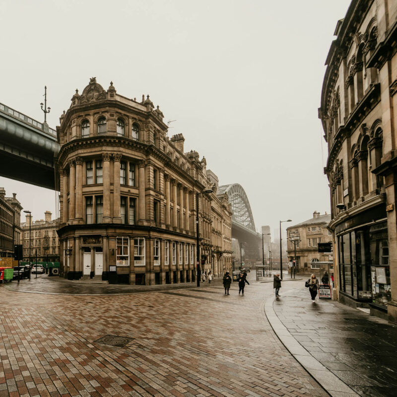 A cobbled street leading down to Newcastle's Tyne Bridge and Quayside on a grey day. A few people walk across the road.