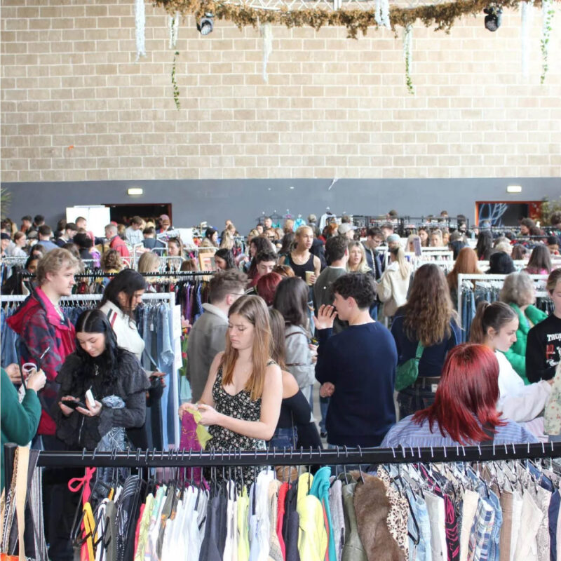 Crowds of people wander down rails of clothes in a large arena. They browse the items on hangers, picking things out to try on and take home.