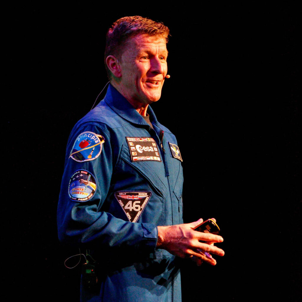 British Astronaut Tim Peake stands against a black background. He wears a blue spacesuit with astronaut badges along the breast and arm. He presses his fingers together and smiles as he looks out at an unseen audience, telling them about his time in space.