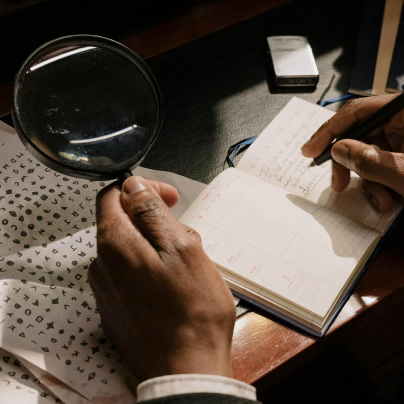 An unseen person holds a magnifying glass over a sheet of coded papers. With their other hand, they hold a pen poised over a notebook.