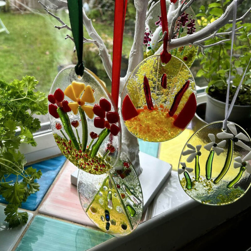 Four glass suncatchers hang from a fake white tree. They are all egg shaped and feature a range of colourful designs depicting flowers, trees and shapes.
