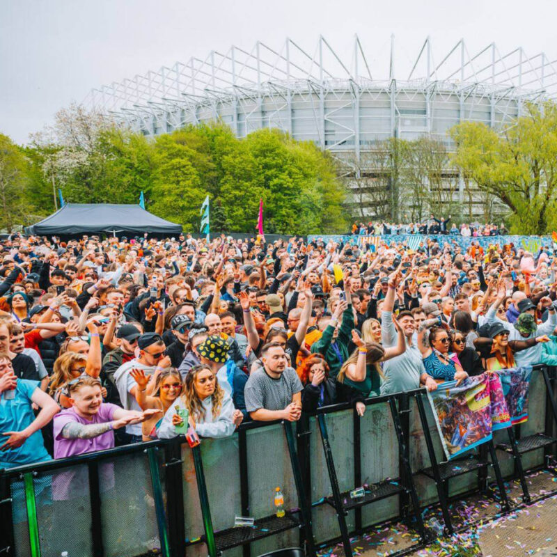 A huge crowd of people dressed in festival attire stand behind a railing facing forward to an unseen stage. Many of them raise their arms in the air and sing along. In the background, the large structure of St James Park stadium can be seen.