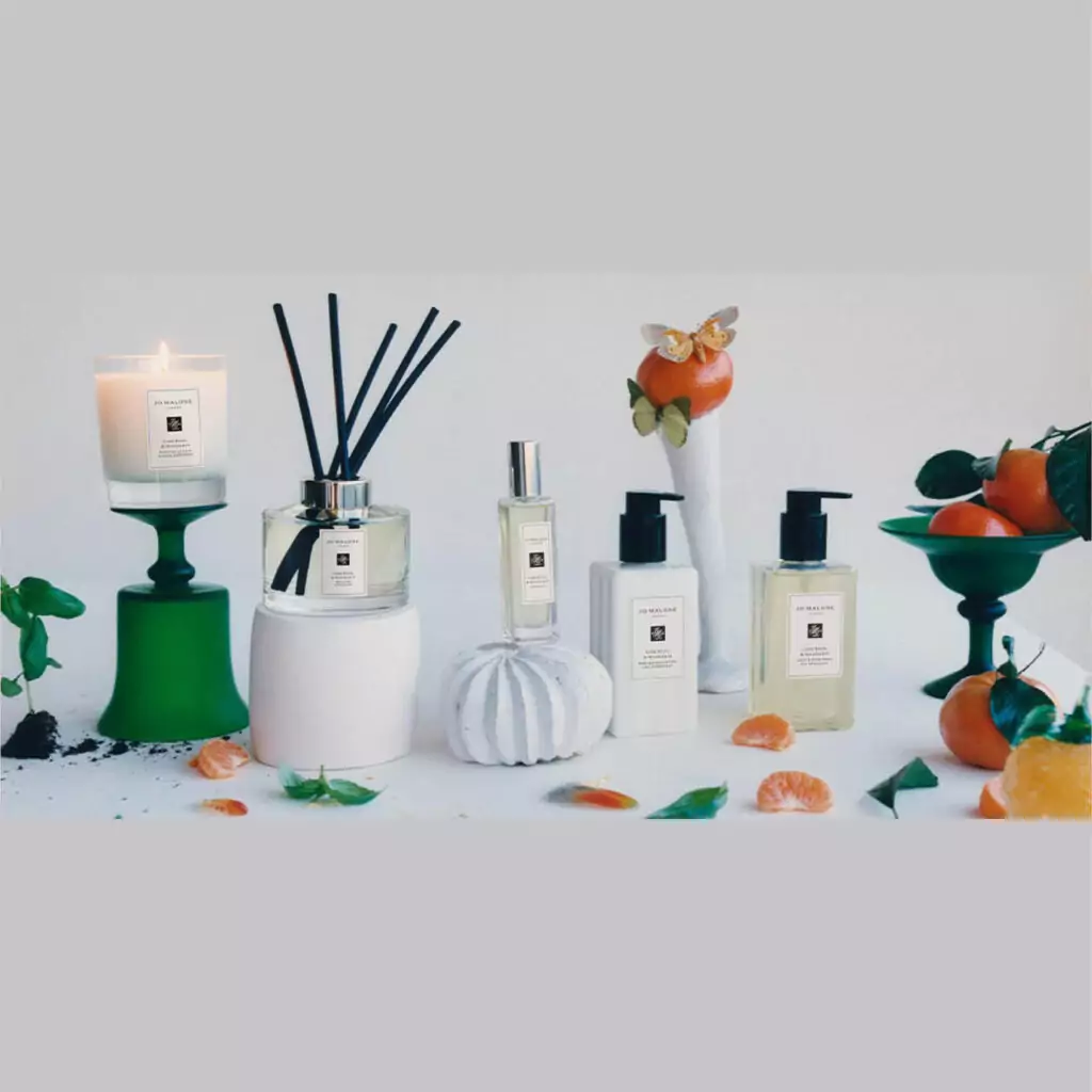 A range of Jo Malone candles. soaps and spray are lined up on a grey surface. Oranges and flower petals surround them.