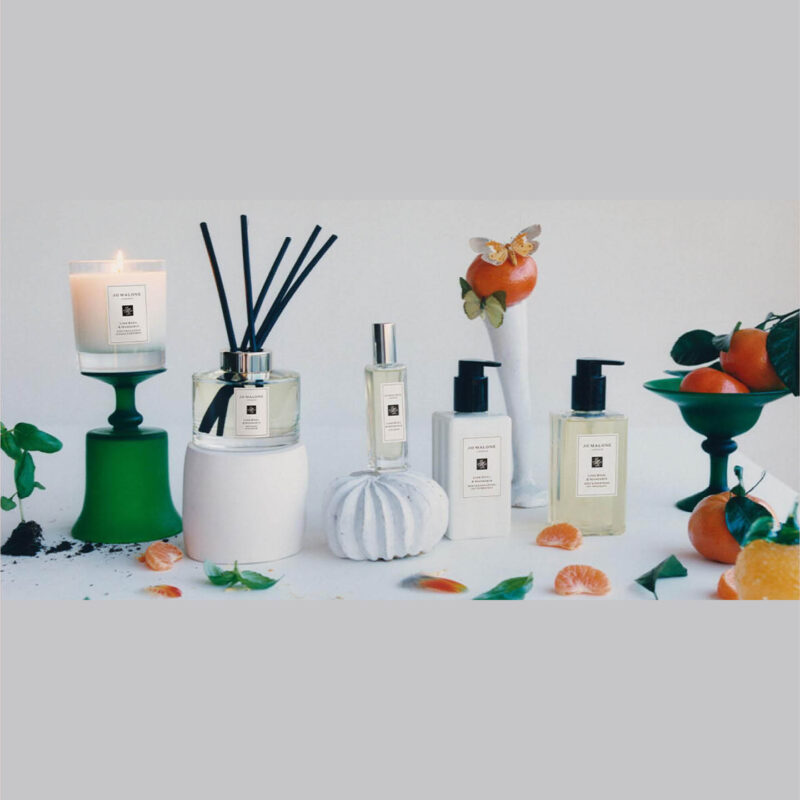 A range of Jo Malone candles. soaps and spray are lined up on a grey surface. Oranges and flower petals surround them.
