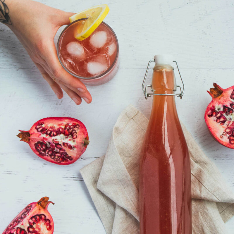 A bottle of red kombucha lies on a white table. Three halved pomegranates are positioned around it. A hand reaches out to grab a glass filled with the kombucha, ice and lemon.