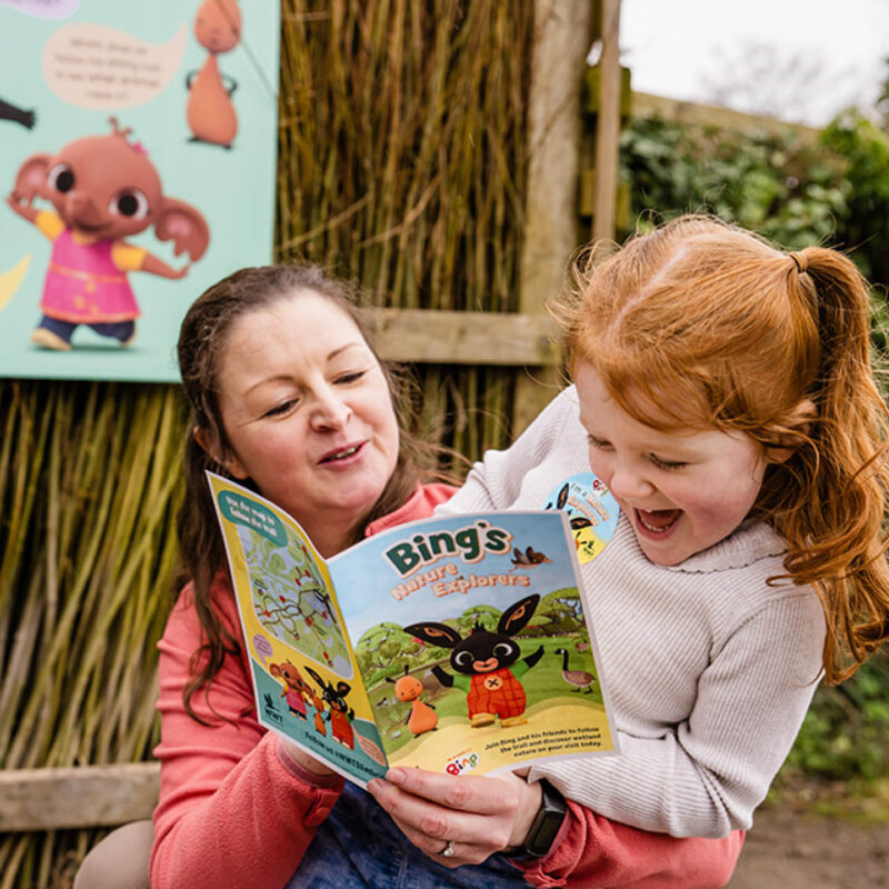 A little girls sits on her mum's lap outside at Washington Wetland Centre. They are reading a Bing book together. The mum points at the page and the little girl laughs happily.