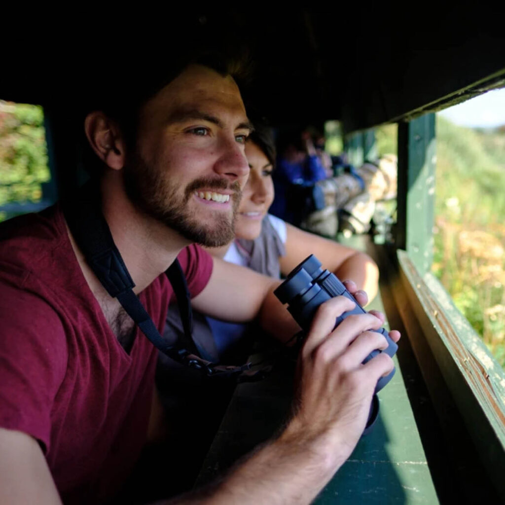 A man stands in a bird hide at Washington Wetlands Centre. He holds a pair of binoculars in his hands and smiles as he looks out the window.