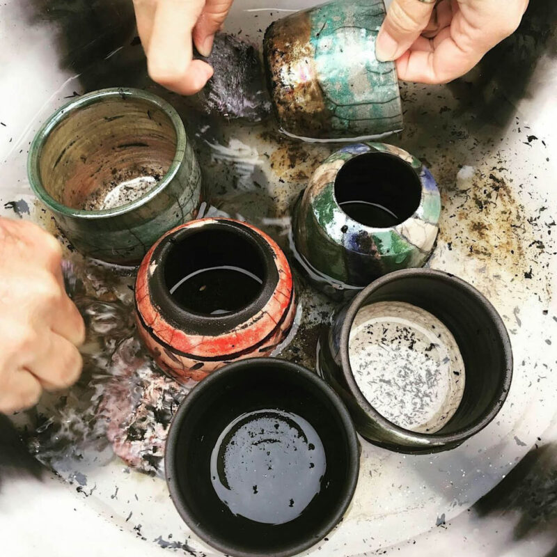 Six newly fired pots sit in a sink, where hands scrub the smoke and soot from their surfaces. The pots' colours already shine through in tones of metallic green, blue, gold and bronze.