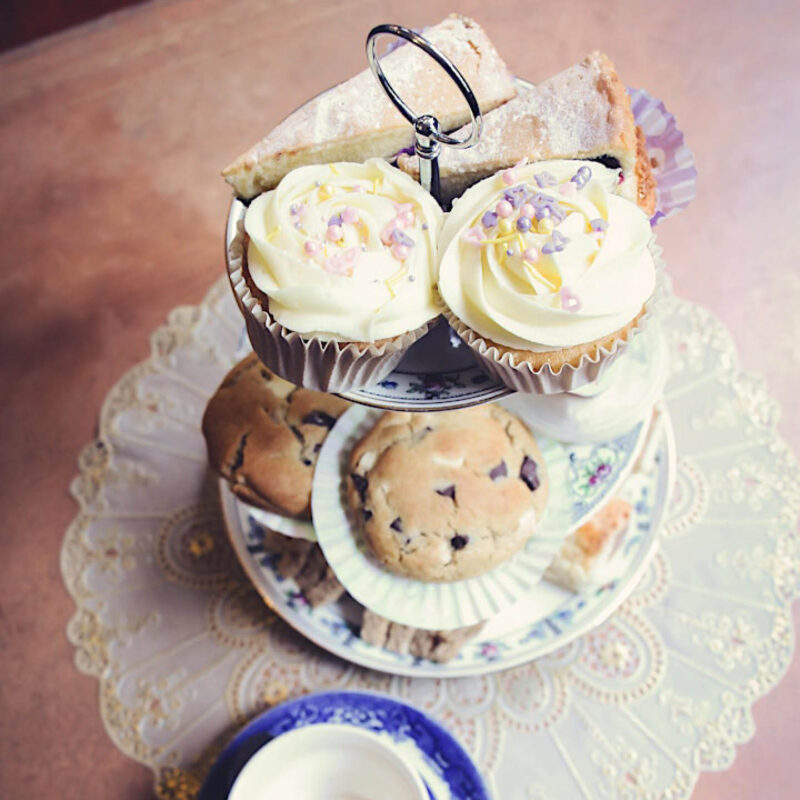 Vanilla frosted tea cups and choc chip cookies are arranged around two tiers of an afternoon tea cake stand.