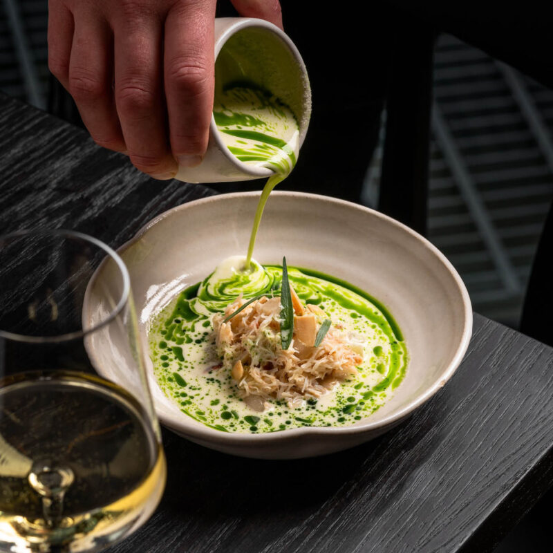 A hand pours a creamy white and green sauce over a plate of crab meat. The sauce swirls prettily around it. A glass of white wine stands in the foreground. It is one of the dishes guests can try with the Spring Tasting Menu at SIX, Baltic.