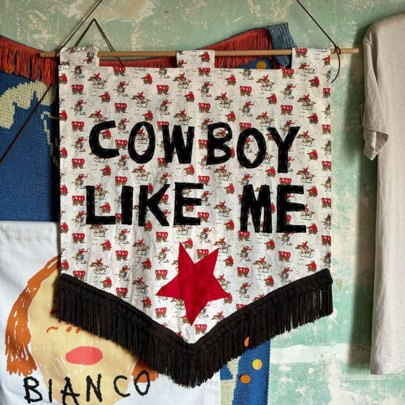 A banner made from cowboy print fabric hanging from a thin pole of wood. On the fabric, the words 'Cowboy like Me' have been stitched on in black fabric. Beneath the text, there is a red star.
