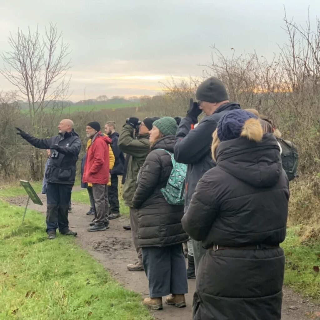 A crowd of people stand along a dirt path at the Washington Wetlands Centre. Fields stretch away in the background. A guide stands at the front of the people, pointing to something in the distance. A couple of people have large cameras raised to their eyes, poised to take a photo of the wildlife.