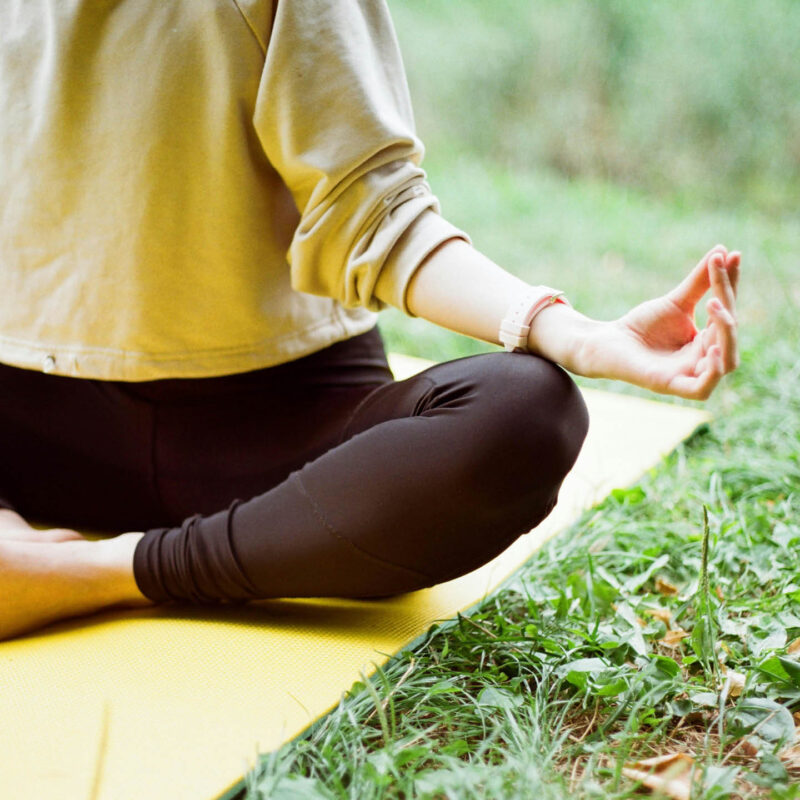 A woman sits on a yellow yoga mat on a patch of grass. Her arms rest on her crossed legs and her fingers pinch together in meditation.