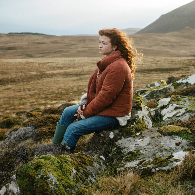 A young red haired woman named Aisling sits on rocks on her home croft, Hoy, before having to move to the nearby town of Stromness. The image is taken by Joanne Coates as part of her exhibition Middle of Somewhere.