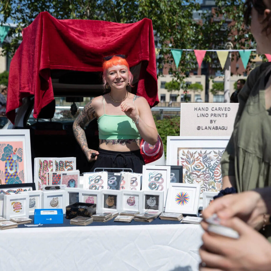 A white woman with dyed orange hair stands in front of her car's open boot. In front of her, there is a white table displaying prints in white frames. The woman laughs as she talks to someone off camera, squinting against the bright sun. She is one of the artists at the BALTIC Car Boot Sale.