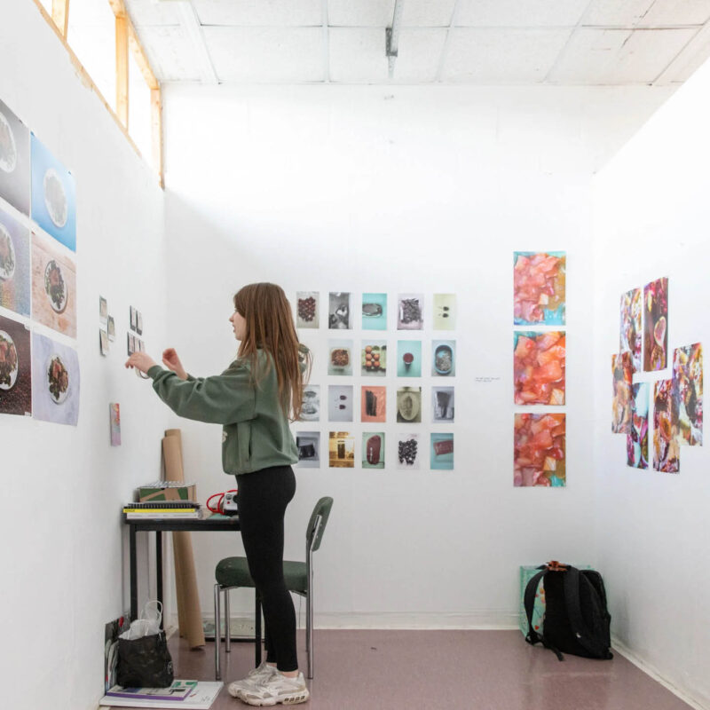 A young woman stands in a small white studio space which has different sized photos and prints on the wall. She positions another piece of art on the wall.