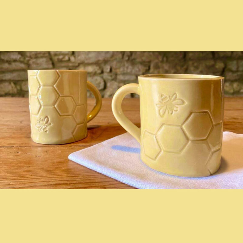 A pair of pale yellow ceramic mugs placed on a wooden surface. They are printed with a honeycomb pattern. A single bee is also stamped into the design.