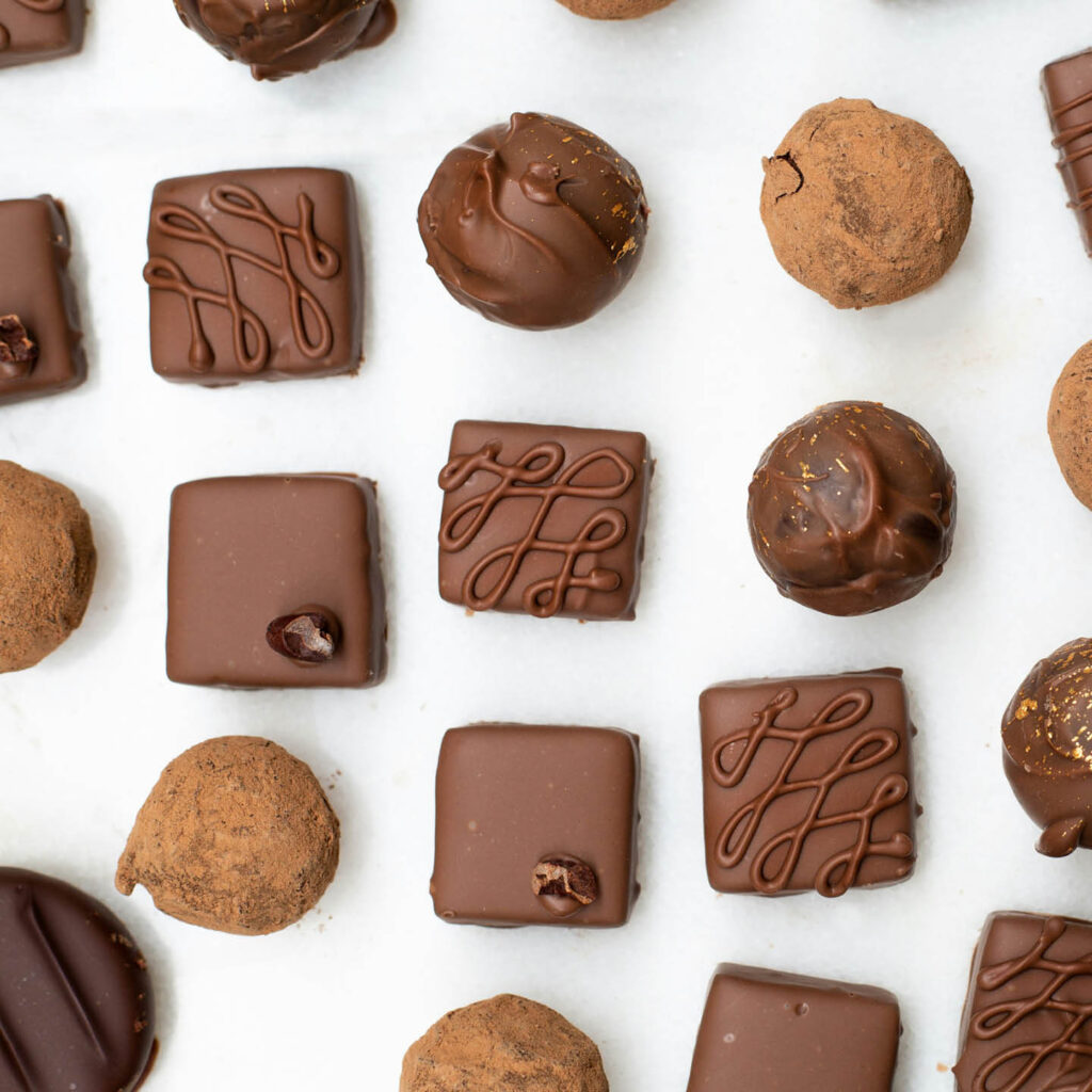 A collection of Belgian truffles are laid out on a white surface. Some of square and smooth, while others are spherical and dusted with cocoa powder or decorated with swirls.