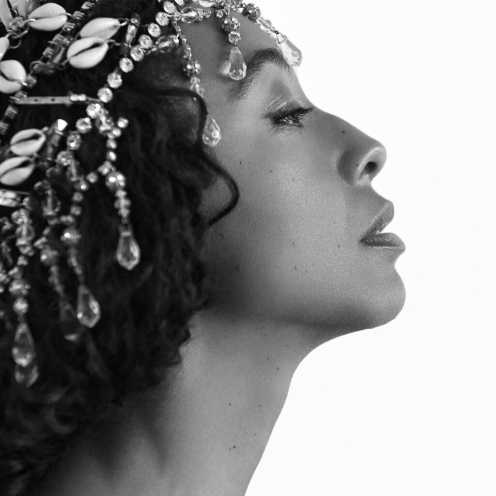 Side profile of Corinne Bailey Rae in black and white. She tilts her chin upwards and looks at something in the distance, an almost pained look on her face. Her black hair is covered with treads of crystals and tiny shells.