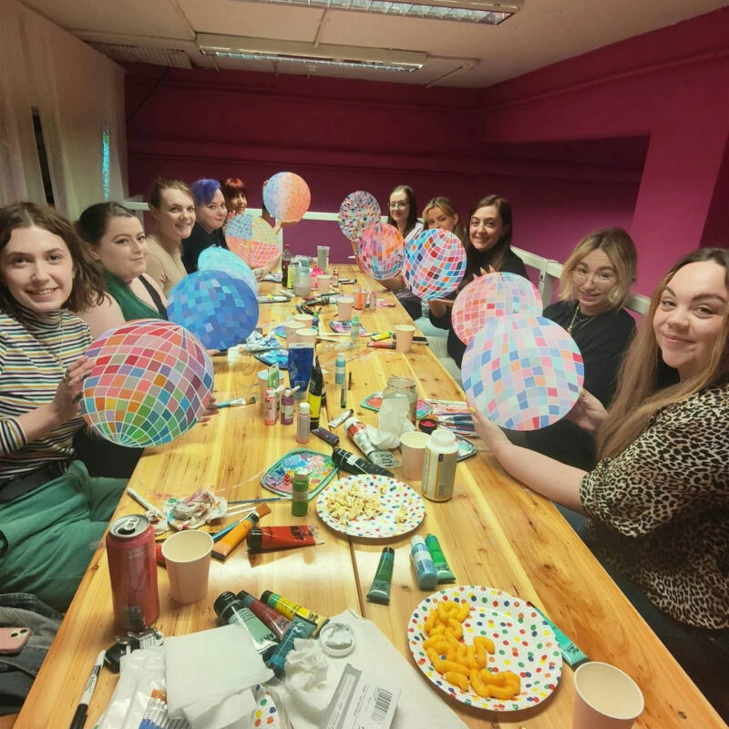 A group of about ten young women line a long wooden table in a pink room. They all hold up a 2D disco ball painted in various colours. They look at the camera and smile. They are taking part in the Eurovision Disco Ball Paint Night at Flaura Rose in Ouseburn.