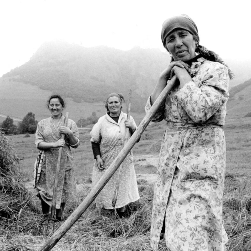 A woman wearing a headscarf and long dress leans on a farming hoe as she looks at the camera. Two women stand further behind her, also wearing long dressing and carrying hoes. They smile at the camera. The image is a black and white photo taken in the Soviet Union by Franki Raffles.
