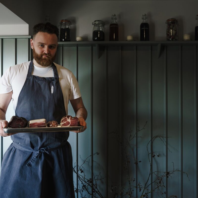 Ian Waller, head chef at Michelin starred Restaurant Pine, stands in front of a blue wood panelled wall. He wears a blue apron and carries a wooden tray holding four cuts of meat. He looks at the camera determinedly.
