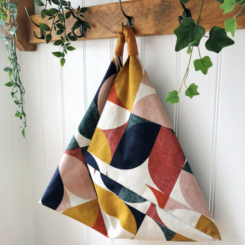 A triangular shaped bag made from colourful geometric patterns hangs from a wooden hook on a wall. It is an origami bag which can be made at the BALTIC Shop's origami bag workshop.