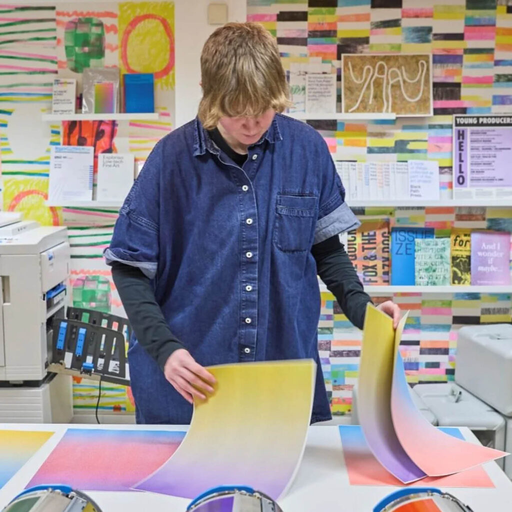 A person wearing an oversized denim shirt stands in a room where colourful prints line the walls. They flick through large sheets of colourful gradient paper.