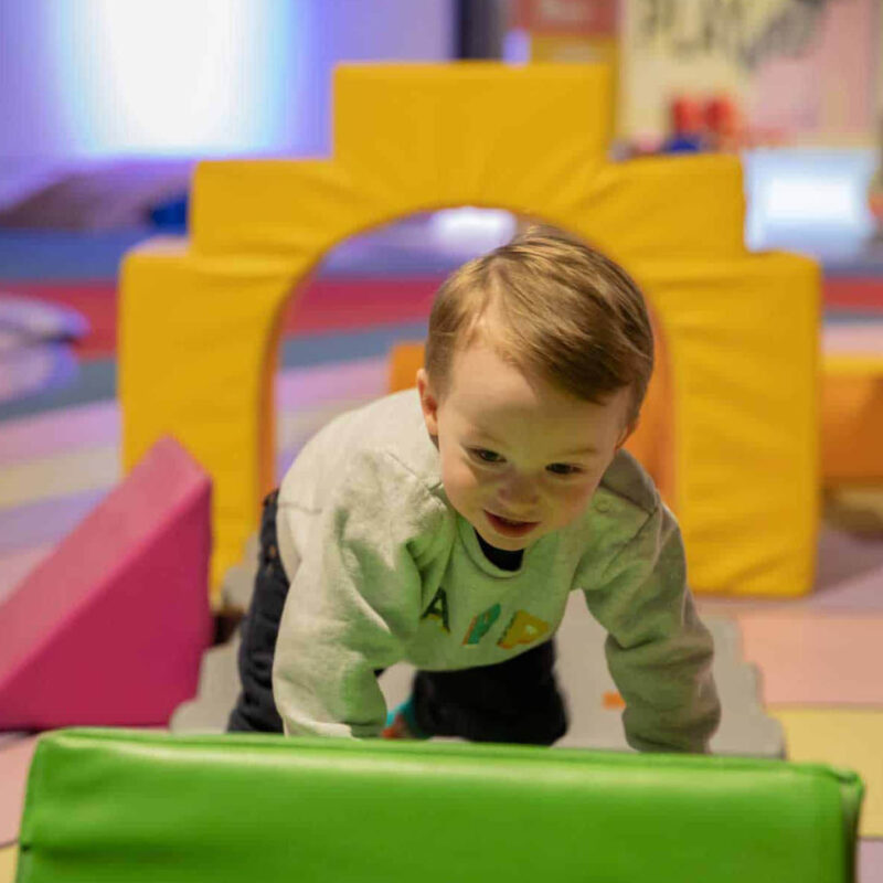 A very little boy with blonde hair clambers up a green squishy triangle. He has a determined smile on his face. Behind him, other foam shapes and arches lie on the floor. The boy is playing at the Life Science Centre during a Pre School Day.