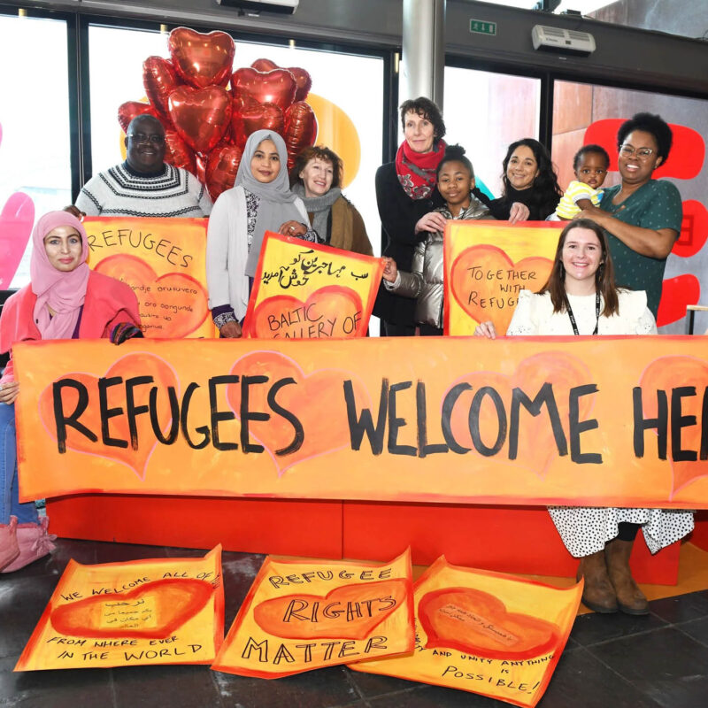 A diverse group of people stand behind a large orange banner which says Refugees Welcome Here. Some of the group hold small posters with hearts and black text painted onto them.