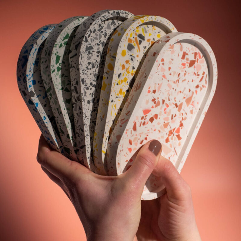 A pair of hands fans out five oval shaped trinket trays. Each one is speckled with a different colour: blue, green, grey, yellow and pink. These trays can be made at the Baltic's Terrazzo Trinket Tray workshop.