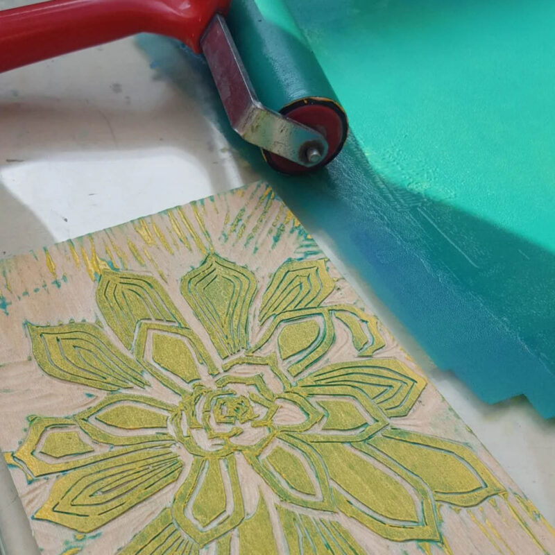 An intricate flower is carved into a large wooden block. It is stained with green paint. Next to it, there is a roller being dipped into a tray of teal paint.
