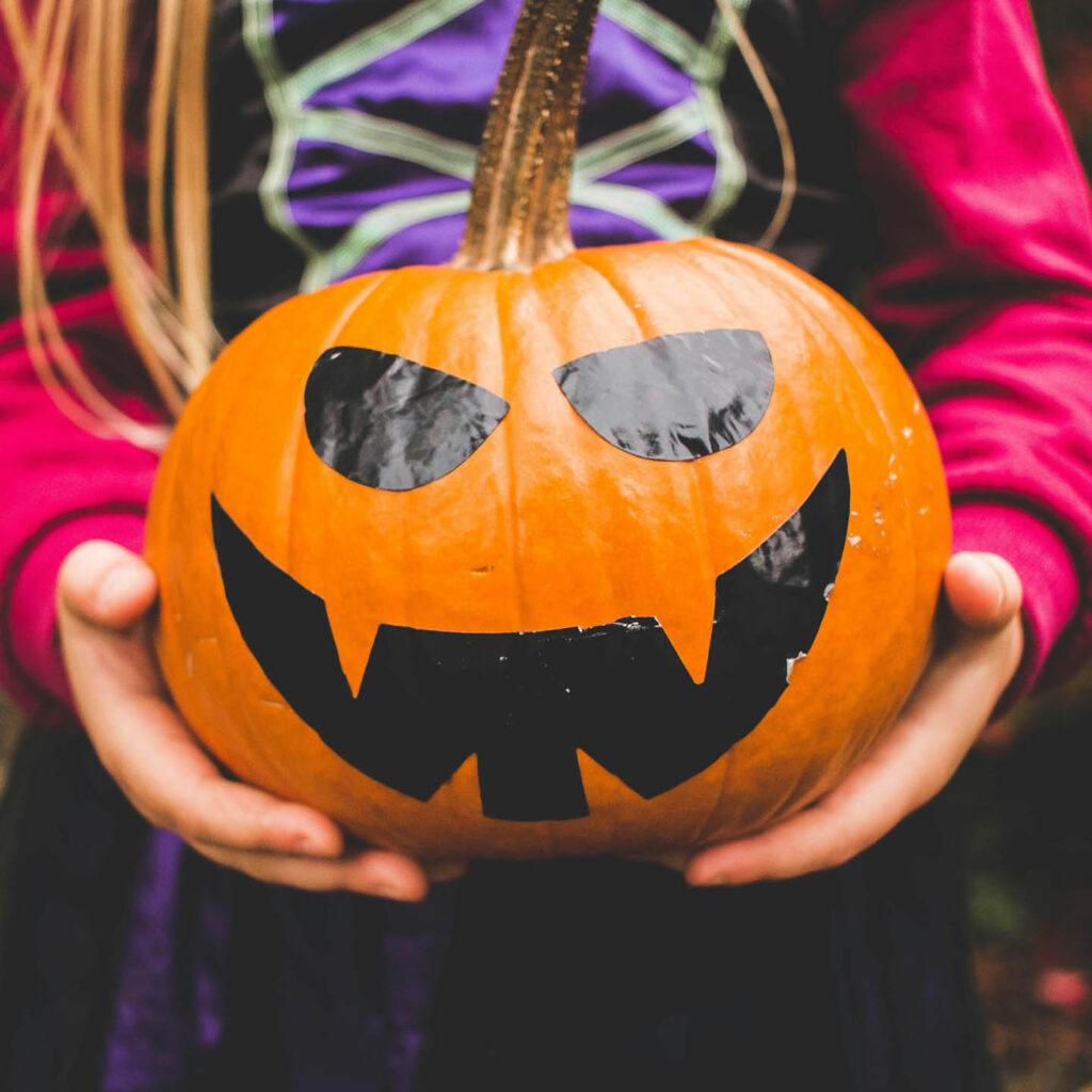 A child wearing a purple, black and pink witch's dress holds a pumpkin. Black eyes and a jagged smile have been drawn onto it.