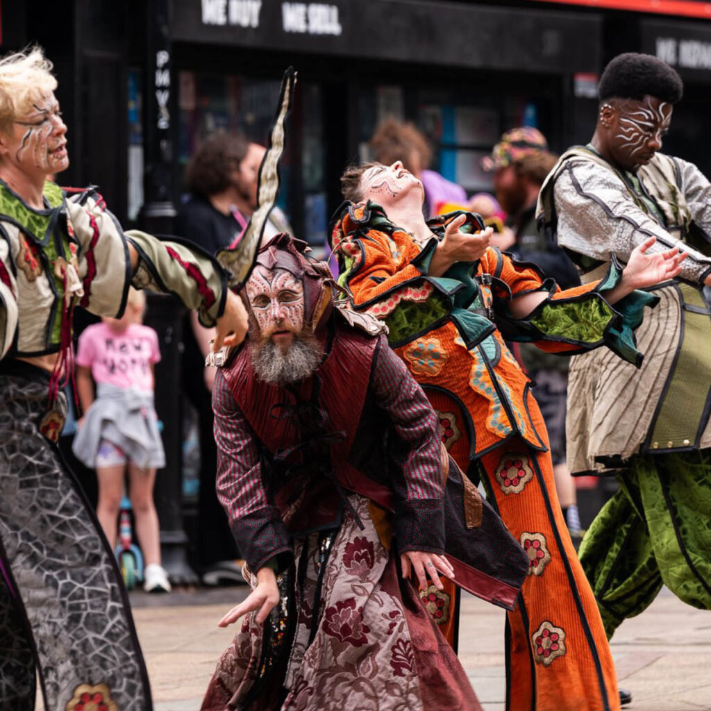 Four people dressed in various coloured printed robes dance in a street in Middlesbrough. Their faces are painted with dots and swirls. They wear pained expressions on their faces, as if they are performing an expressive dance.