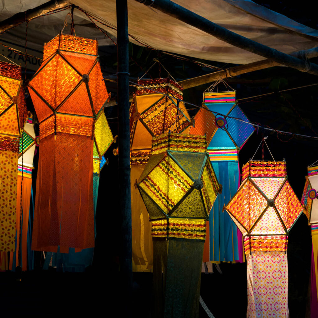 A cluster of different coloured paper lamps with long trailing tassels hang from a ceiling. They are illuminated on the inside, highlighting their beautiful designs for Diwali.