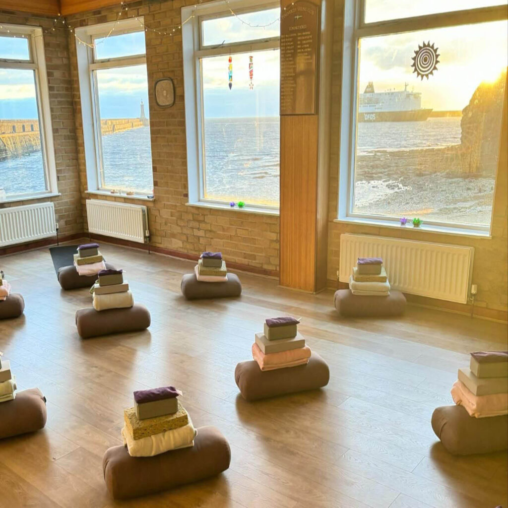 Neat stacks of blankets, blocks and towels are positioned on a wooden floor inside the Jasmine Yoga Studio, Tynemouth. Floor to ceiling windows line the walls, letting in golden evening sunlight and showing off views of the ocean.